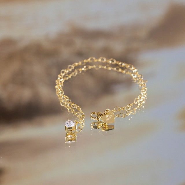 Endloses PURELEI Endless Love Armband in 18K Gold mit Zirkonia-Herzanhänger, elegant auf spiegelnder Oberfläche.
