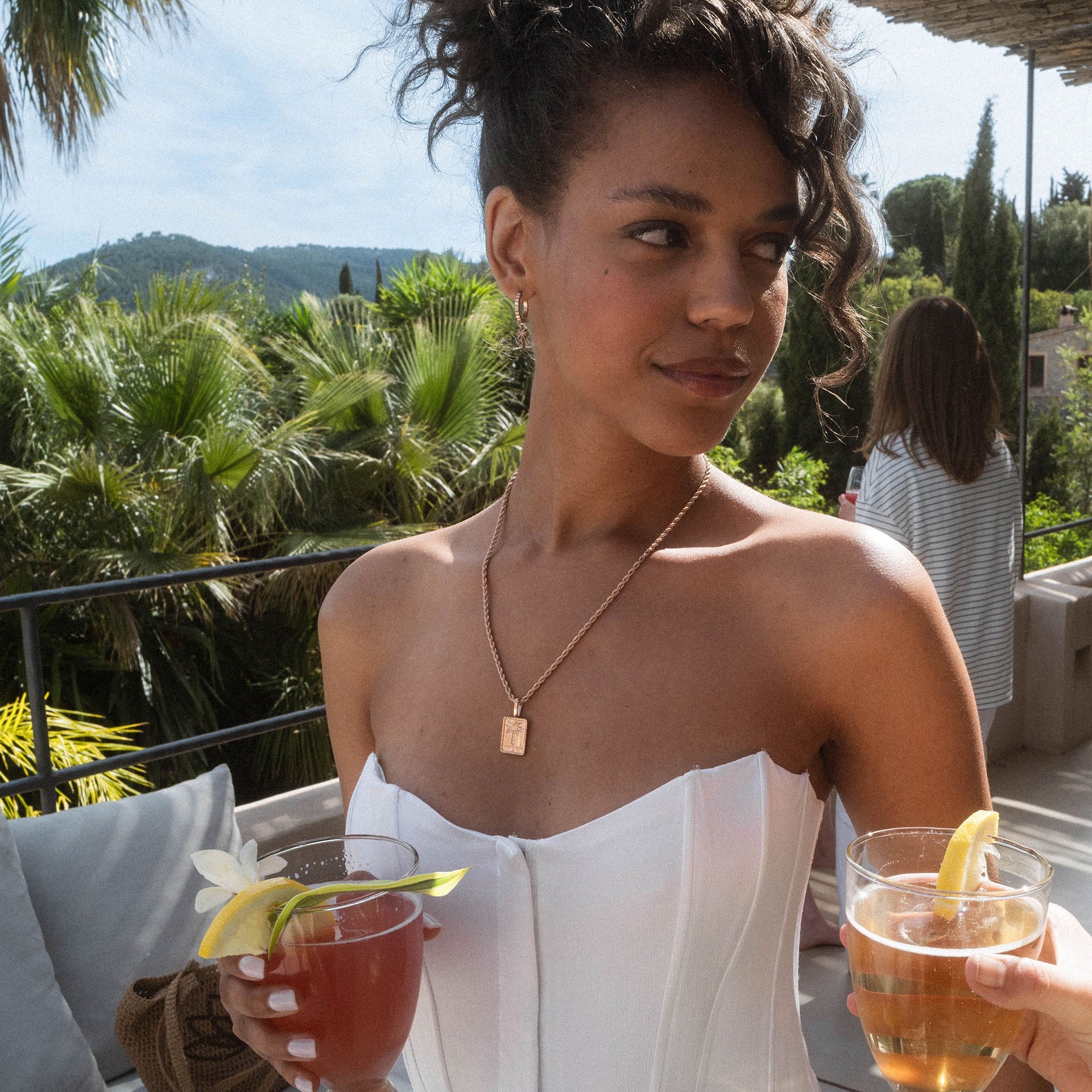 Goldene PURELEI Kette mit rechteckigem Anhänger, elegant und feminin, umgeben von tropischer Atmosphäre.