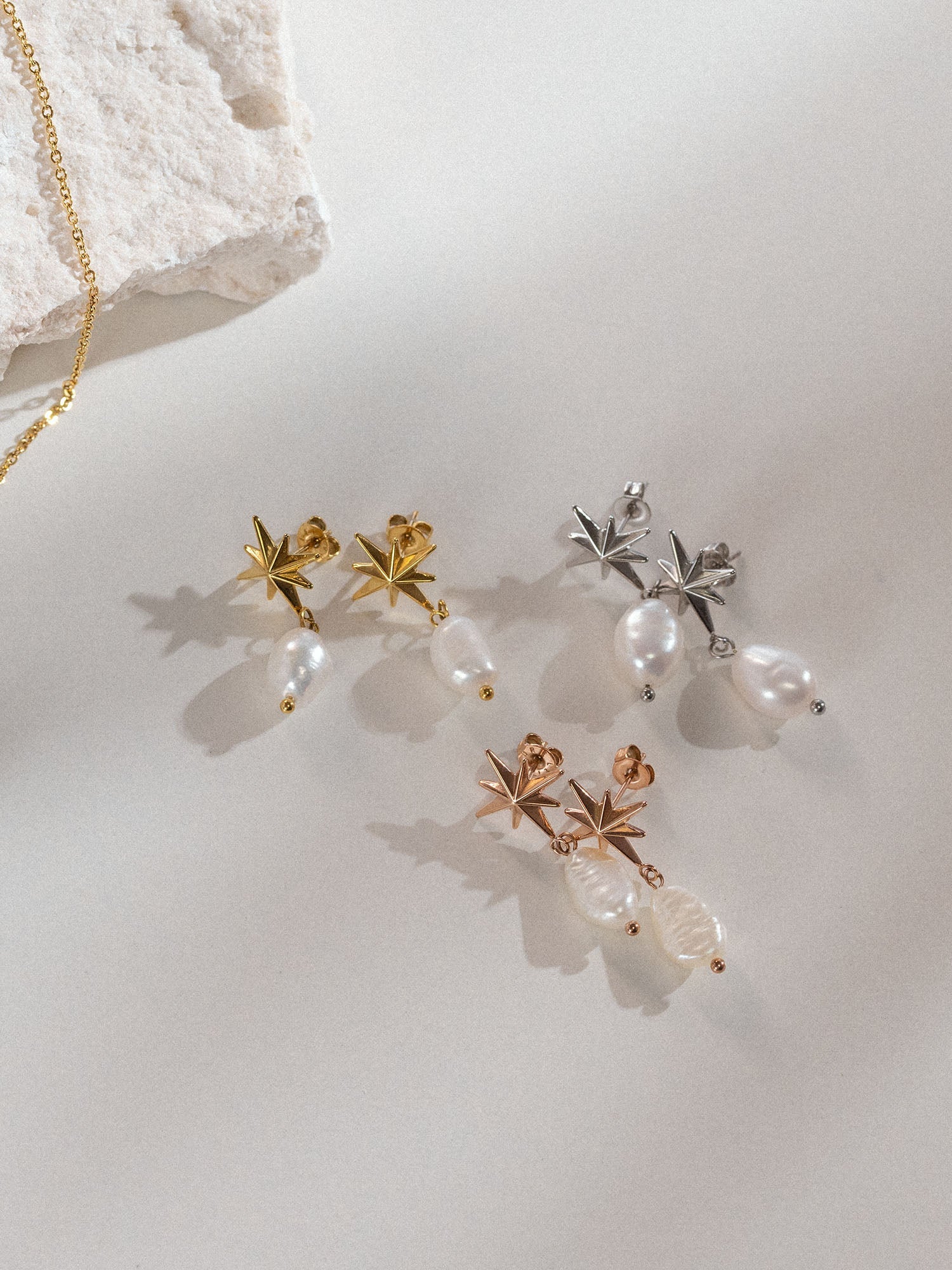 Mahina Club Windrose Ohrringe in Gold, Silber und Roségold mit schimmernder Süßwasserperle, elegant inszeniert auf hellem Untergrund.