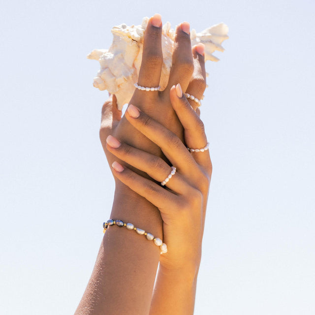 Zarte PURELEI Ringe und Armbänder mit Perlen, in warmem Licht gehalten, von Händen mit Muschel.