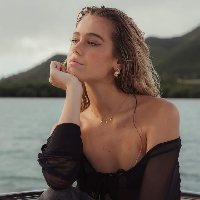 Glänzende PURELEI Glowing Ohrringe in Gold, breites Design, getragen mit tropischer Bergkulisse im Hintergrund.