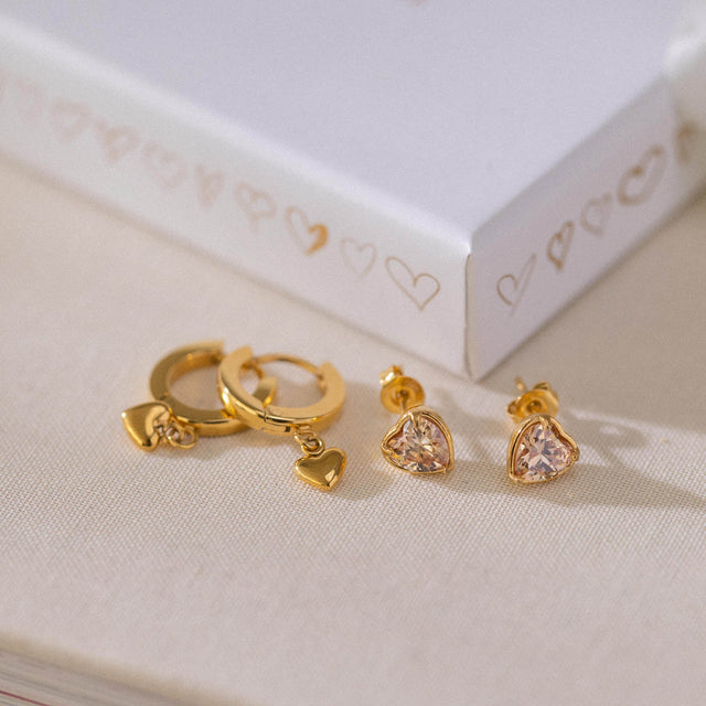 Goldene Ohrringe mit Herzanhängern und funkelnden Steinen von PURELEI auf hellem Stoff.