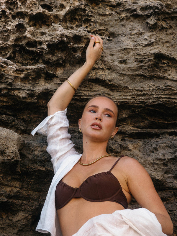 Elegante, braune Bikini-Oberteil mit PURELEI Schmuck, vor rauem Felsen, tropische Stimmung.