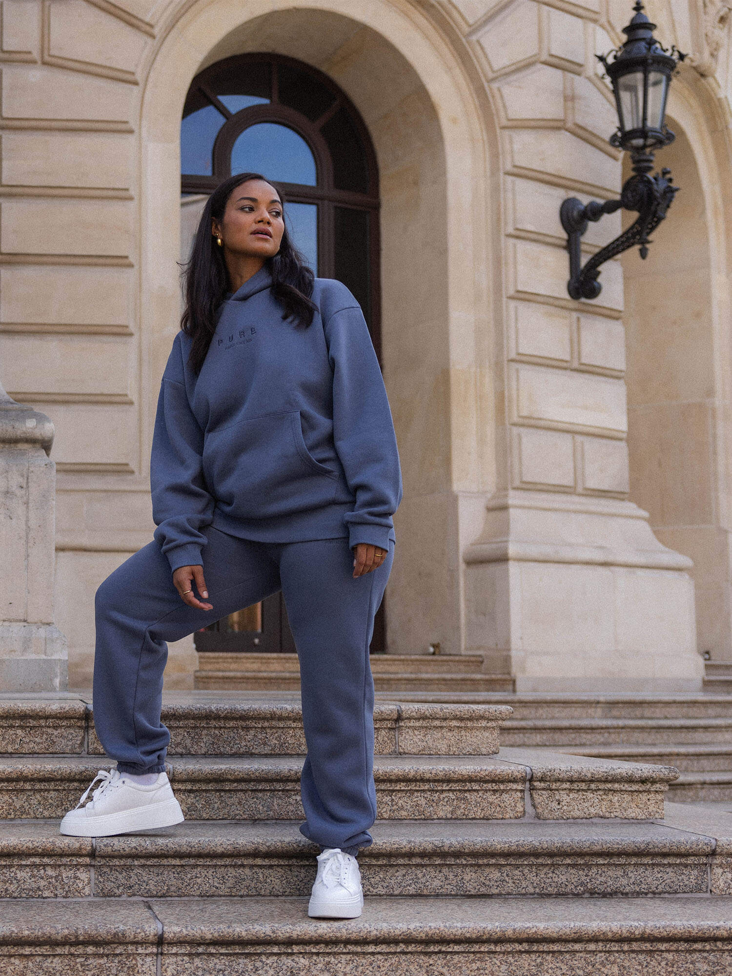 Bequemer, blauer Hoodie und Jogginghose von PURELEI, lässig vor eleganter Architektur.