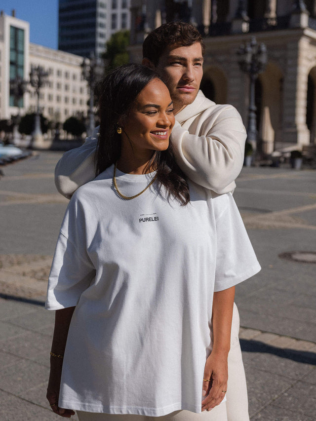 Zwei Personen in weißen PURELEI T-Shirts, lächelnd in urbaner Umgebung.