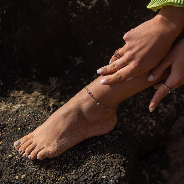 Zartes PURELEI Fußarmband aus Roségold mit Herzanhänger, getragen auf sandiger Oberfläche.