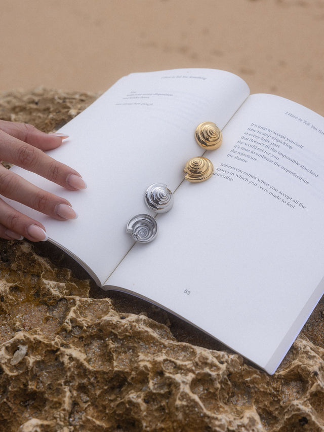 PURELEI Nautical Big Ohrringe in Gold und Silber mit Muschelspirale auf offenem Buch am Felsen, elegant maritim.