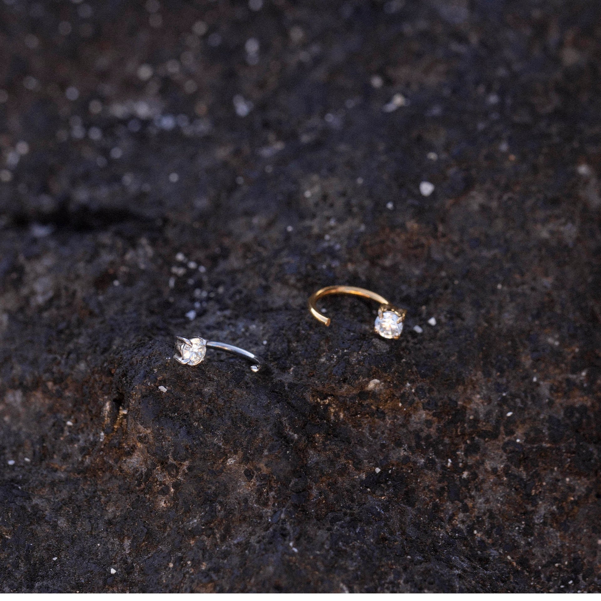 Highlight Piercing von PURELEI, gold- und silberfarbene Edelstahlringe mit funkelndem Zirkonia auf dunklem Lavagestein.