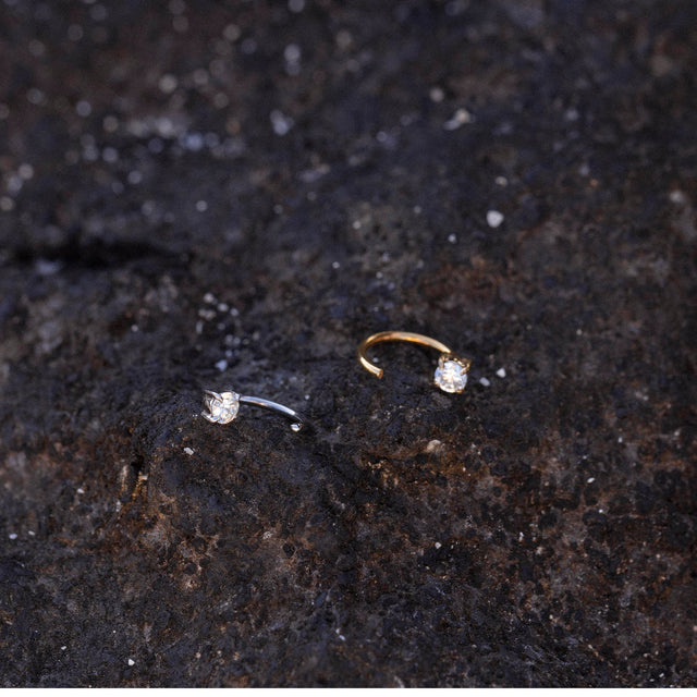 Highlight Piercing von PURELEI, gold- und silberfarbene Edelstahlringe mit funkelndem Zirkonia auf dunklem Lavagestein.