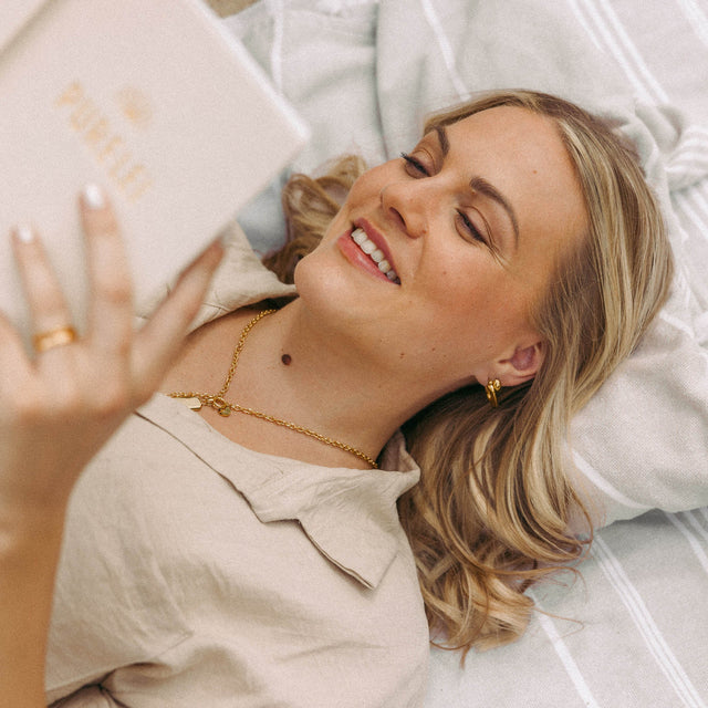 Frau mit PURELEI Schmuck, lächelnd und lesend, in sanften Erdtönen auf weichem Stoff.