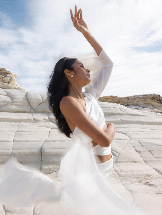 Elegantes PURELEI Leo Letter Charm in Gold mit modernem Design, getragen vor heller Felslandschaft, sanfte Leichtigkeit.