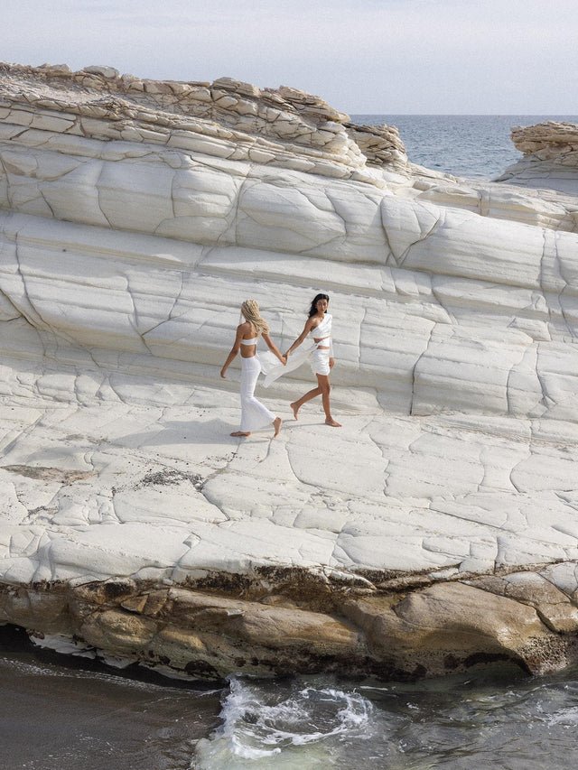 Zwei Frauen laufen barfuß am Strand, beide tragen PURELEI Schmuck, leichte Sommerkleidung, natürliche Eleganz, Meereshintergrund