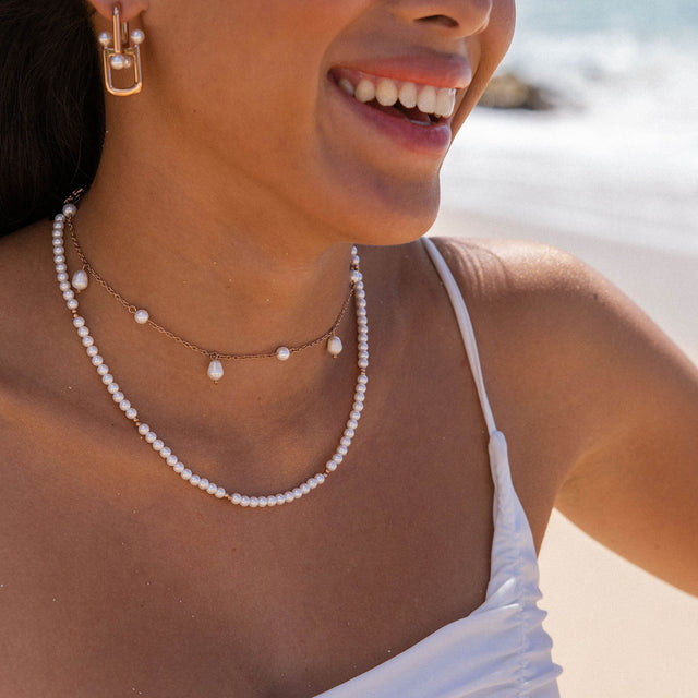 Zarte PURELEI Perlenkette und Ohrringe, elegant am Strand getragen, in warmem Licht.