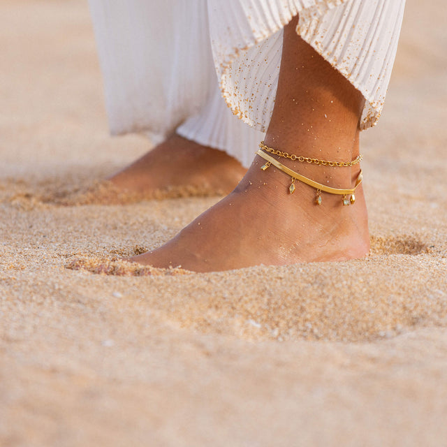 Goldenes Fußarmband mit Anhängern, getragen im Sand, von PURELEI.