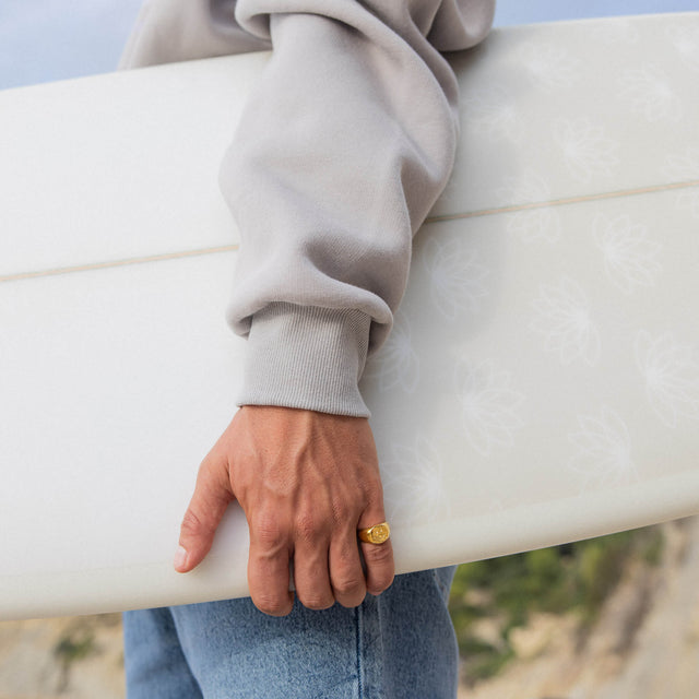 Helle Surfbrett mit floralem Design, gehalten von einer Person in grauem Sweatshirt, PURELEI.